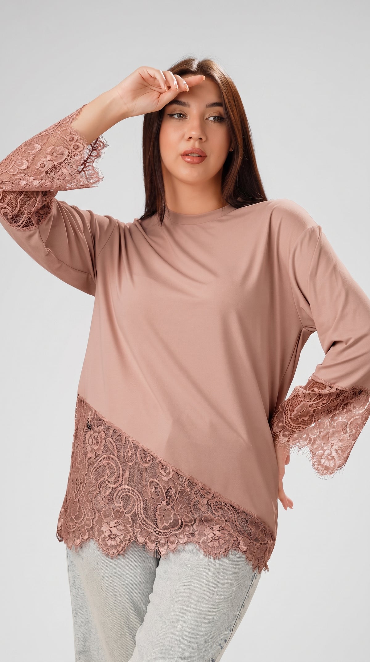 A woman modeling a dusty rose long-sleeve top with lace trim on the sleeves and hem, paired with light gray jeans, against a solid light gray background.