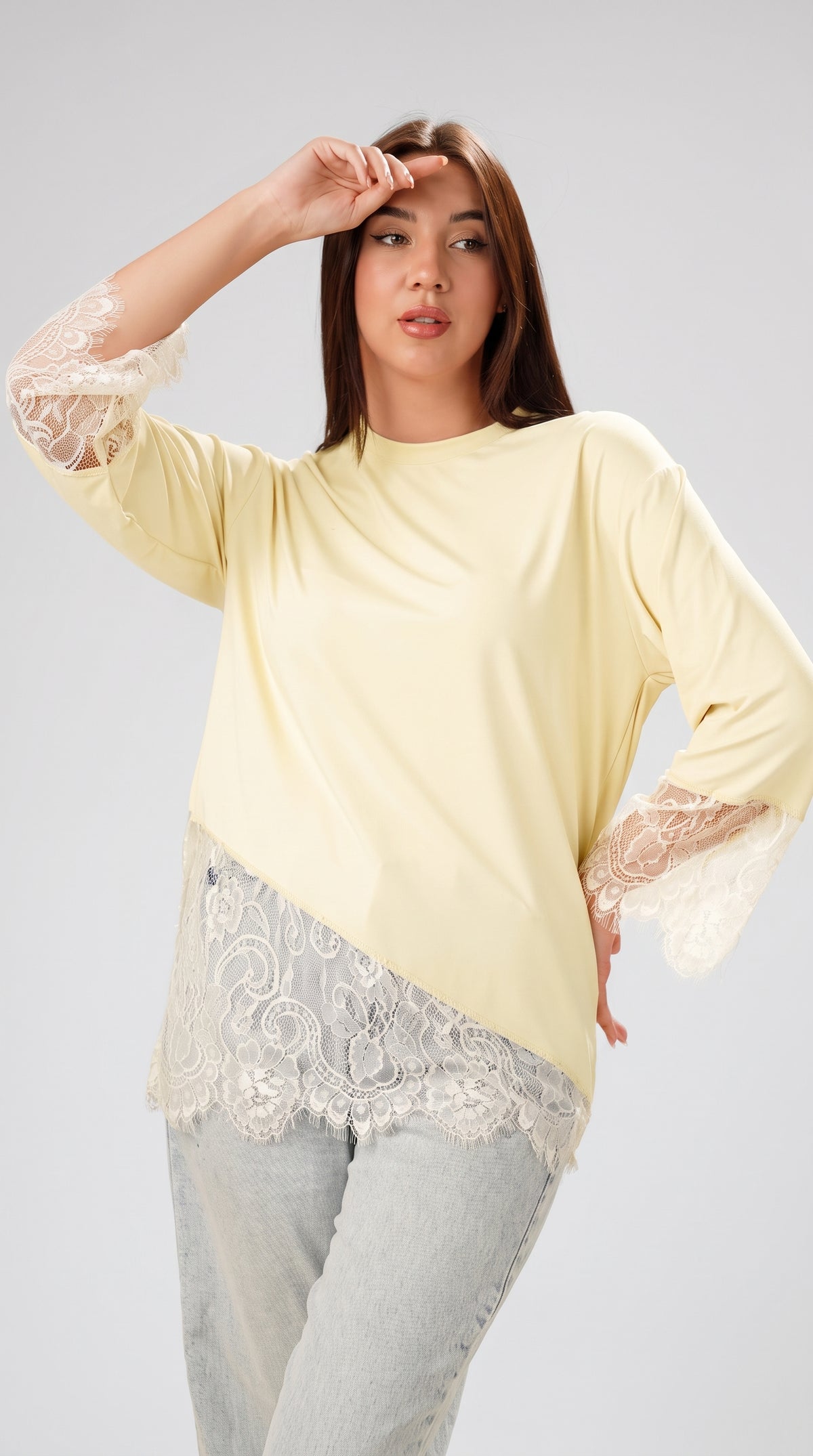 A woman modeling a pale yellow asymmetrical long-sleeve top with white lace trim on the sleeves and hem, paired with light gray jeans, against a light gray studio background.