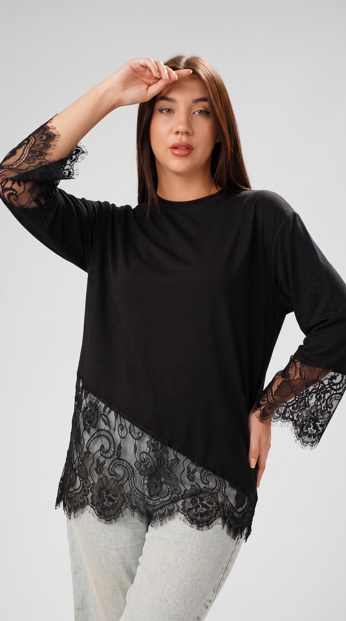A woman modeling a black long-sleeve top with sheer black lace trim at the cuffs and hem, paired with light gray jeans, against a light gray studio background.