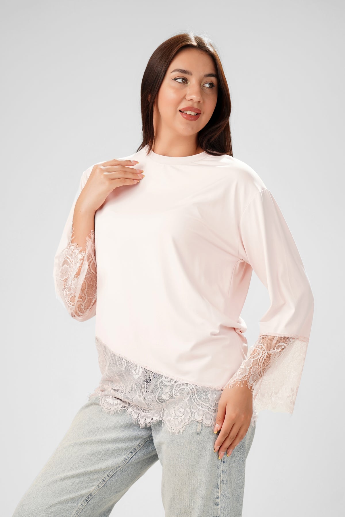 A woman modeling a light pink long-sleeve top with sheer lace bell sleeves and a matching lace hem detail, paired with light-wash jeans, against a solid light gray studio background.
