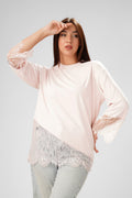 A woman modeling a light pink long-sleeve top with white lace trim on the sleeves and hem, paired with light-wash jeans, against a solid light gray background.
