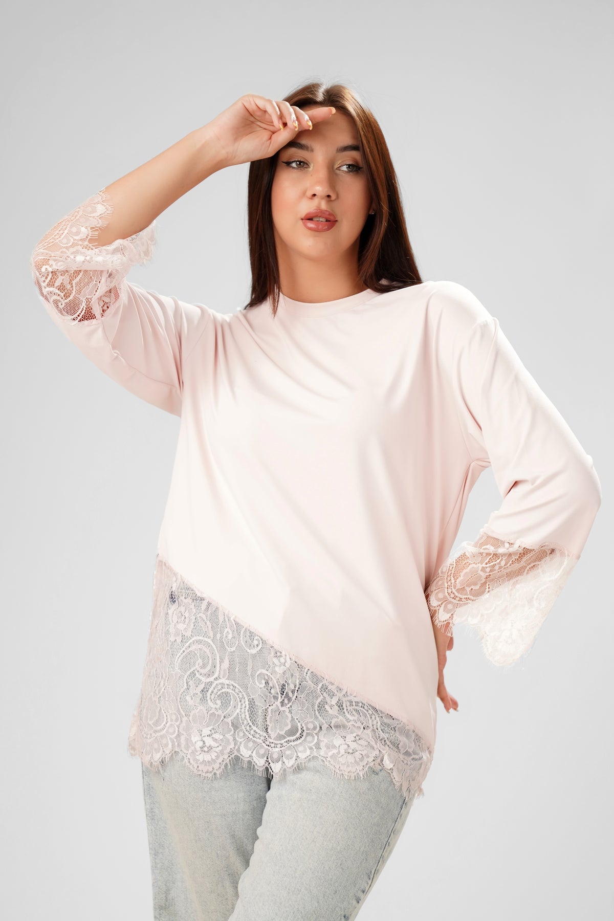 A woman modeling a light pink long-sleeve top with white lace trim on the sleeves and hem, paired with light-wash jeans, against a solid light gray background.