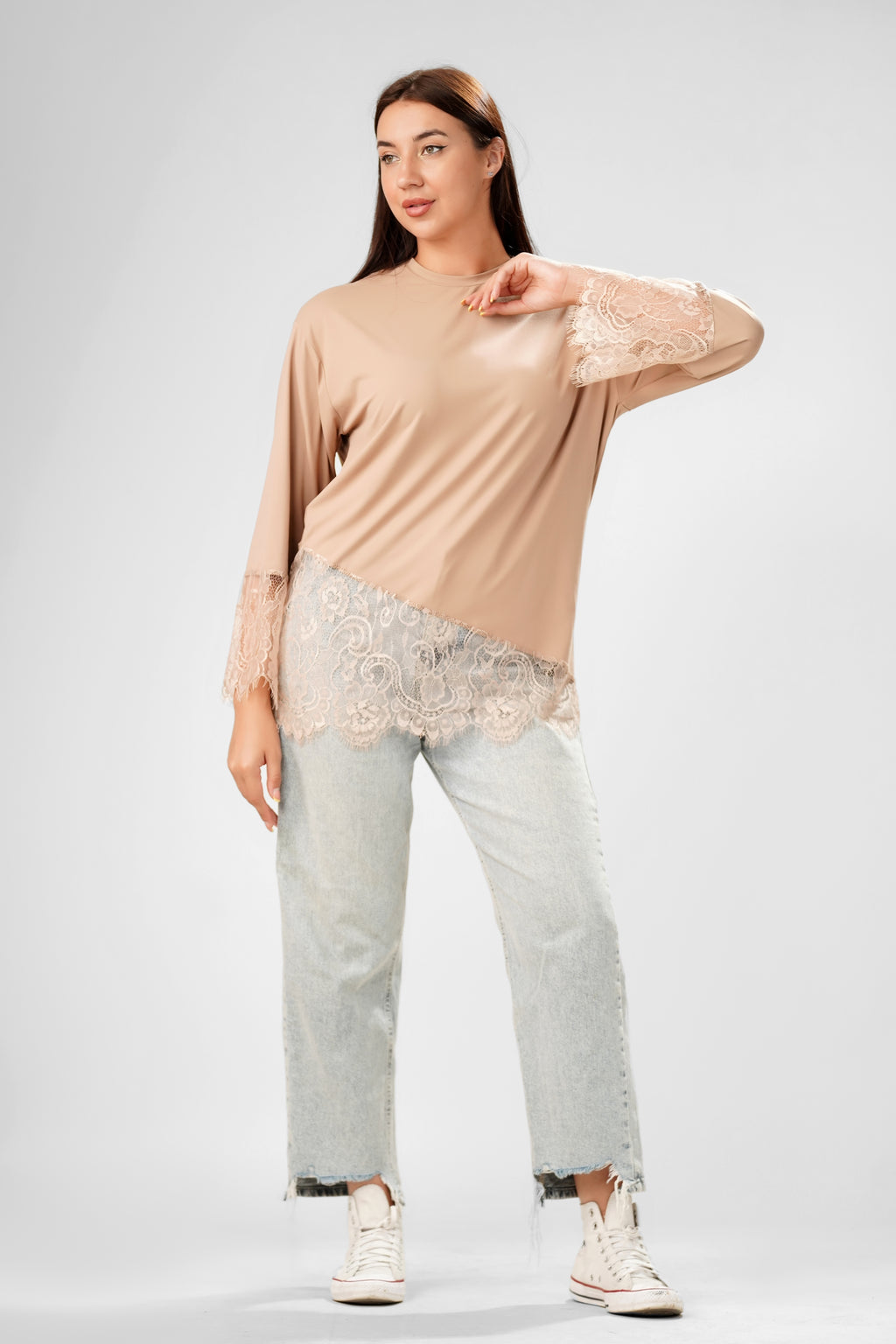 A woman modeling a beige long-sleeve top with lace trim on the sleeves and hem, paired with light-wash distressed jeans and white sneakers, against a light gray studio background.