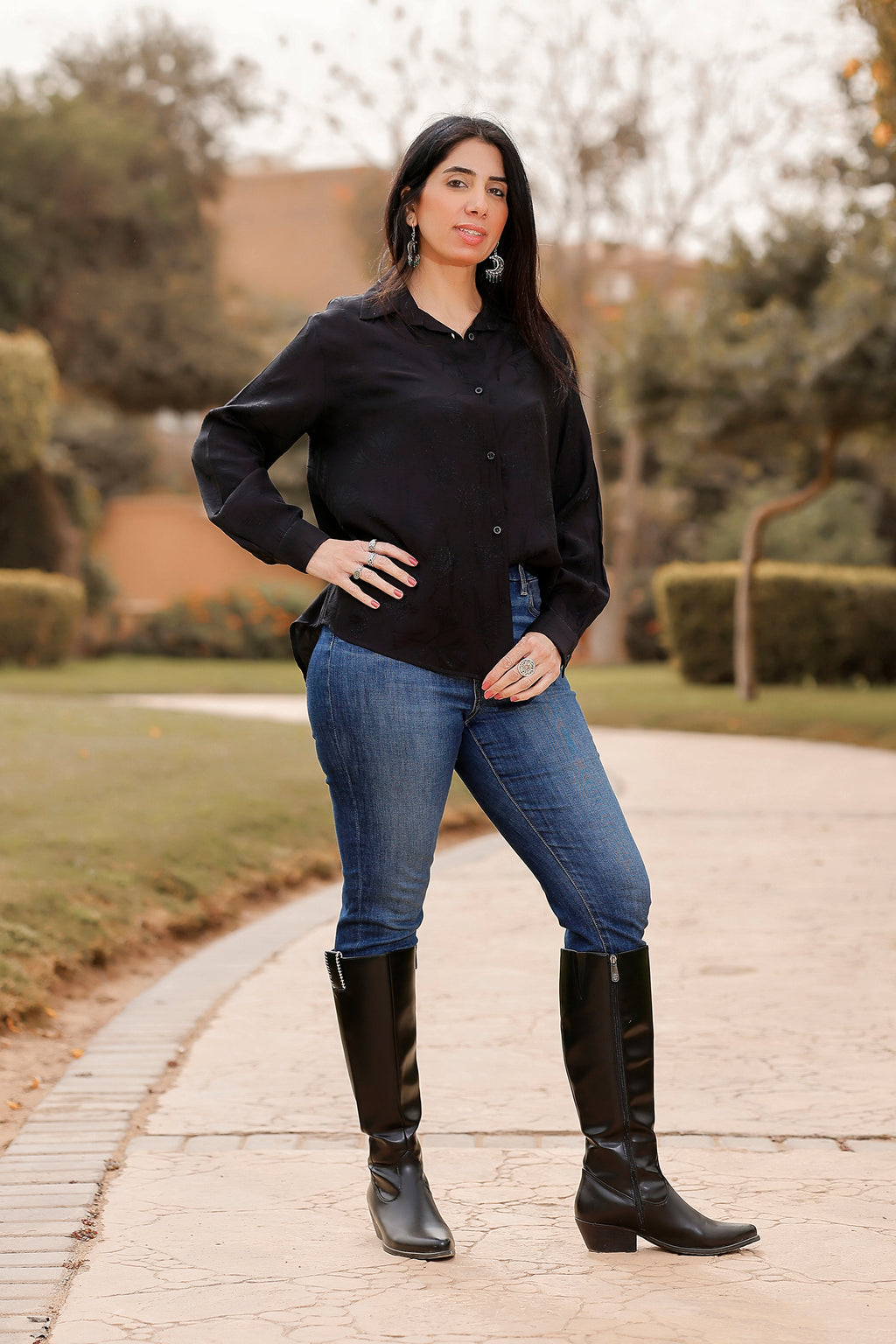 Woman wearing a black shirt, blue jeans, and black knee-high boots standing on a paved path.