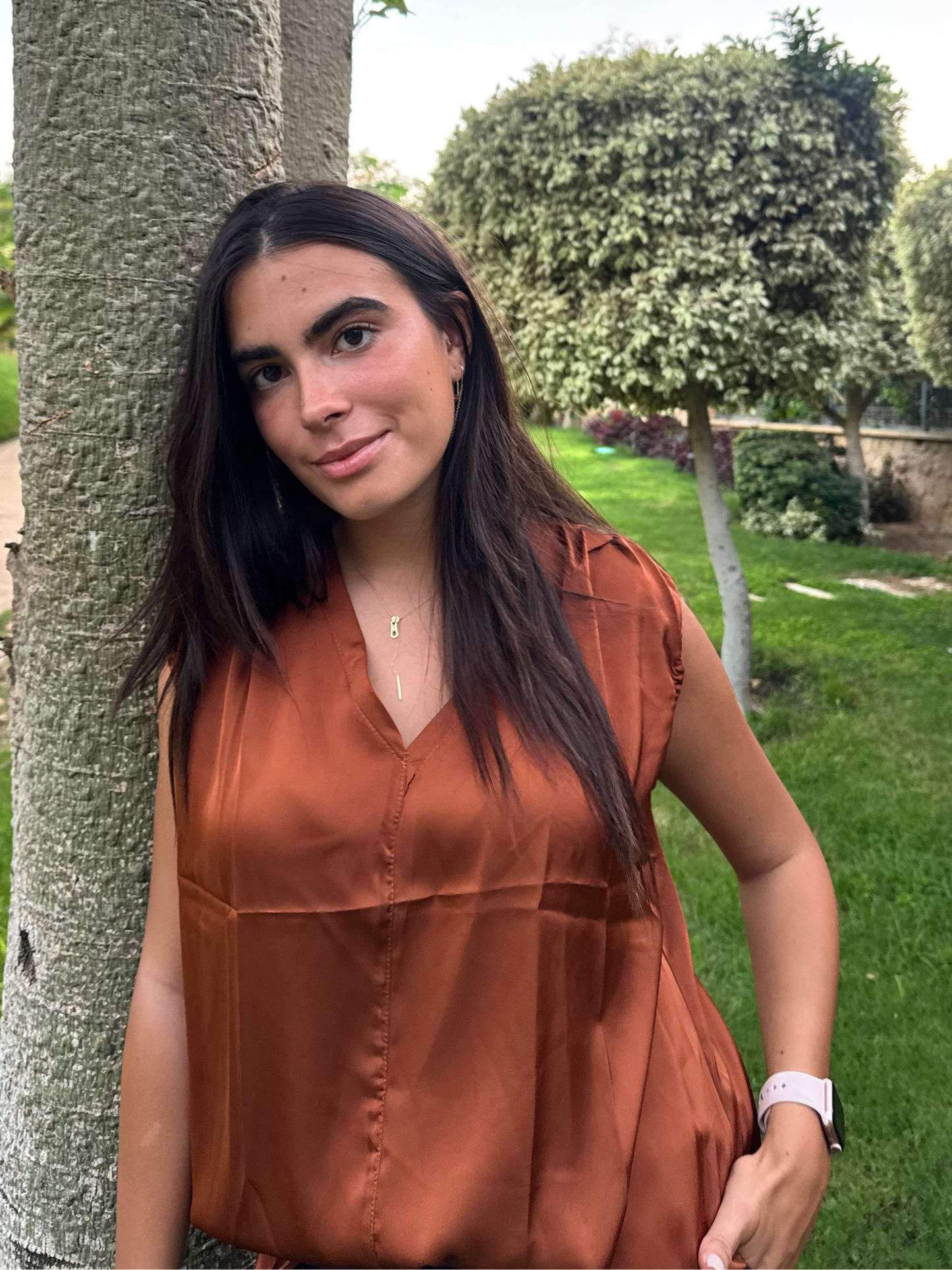 Woman in a rust-colored blouse standing next to a tree in a park.