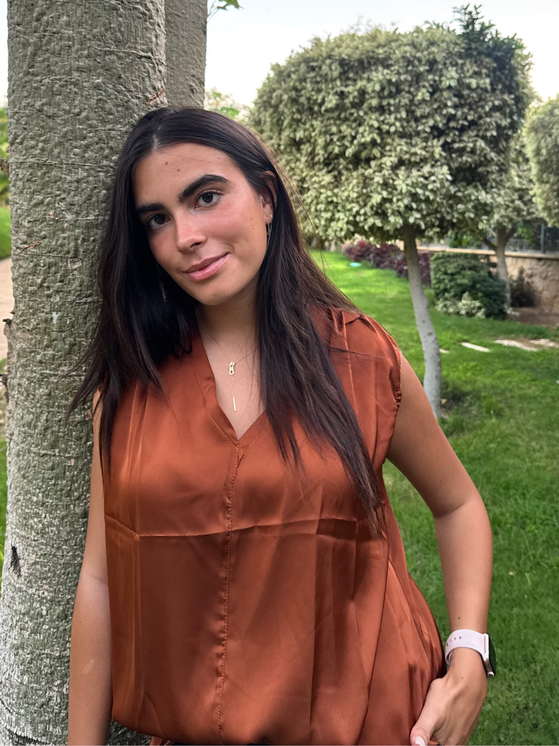 Woman in a rust-colored blouse standing next to a tree in a park.