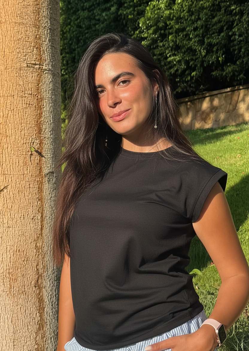 A woman with long dark hair stands outdoors next to a tree trunk, wearing a black short-sleeve crewneck t-shirt and light blue and white striped trousers.