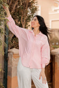 Woman wearing a pink blouse and white pants standing outdoors with plants and a building in the background.