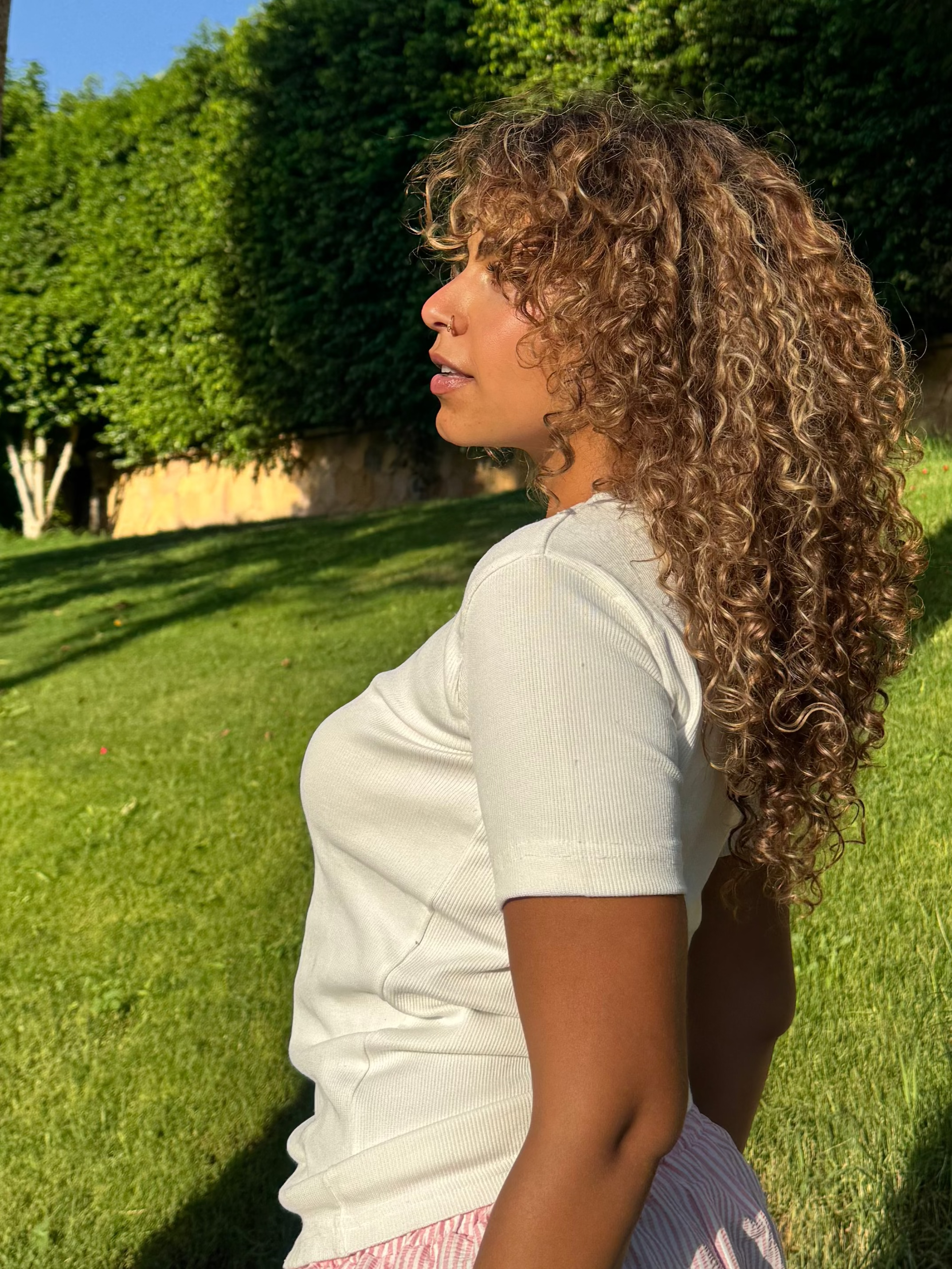 Woman with curly hair standing outdoors on a grassy area with trees in the background