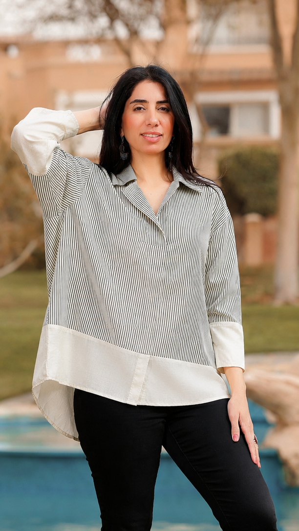 A woman stands outdoors in front of a pool and landscaped background, wearing a light gray and white color-blocked long-sleeve blouse, black pants, and brown knee-high boots.