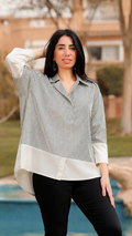 A woman stands outdoors in front of a pool and landscaped background, wearing a light gray and white color-blocked long-sleeve blouse, black pants, and brown knee-high boots.