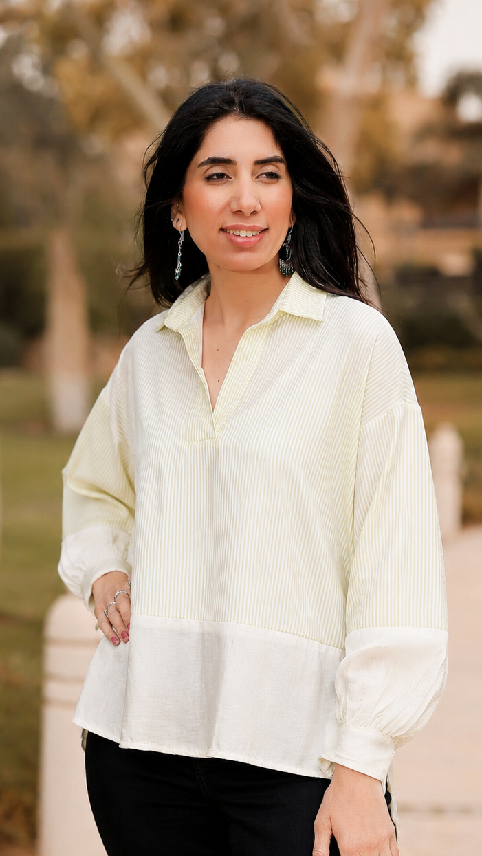 A woman wearing a cream and white long-sleeve collared blouse with vertical texture on the upper portion and a solid white lower panel, paired with black pants, standing outdoors on a paved path with trees in the background.